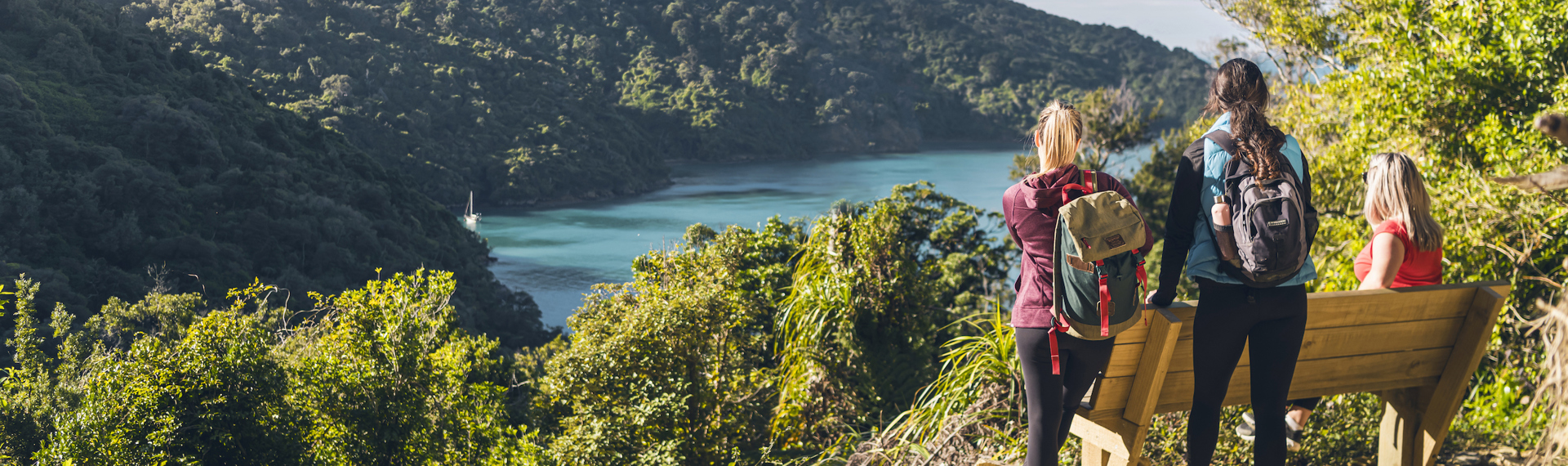 Three people at a bench and viewpoint on the northern Queen Charlotte Track, overlooking Ship Cove/Meretoto in the Marlborough Sounds, New Zealand.