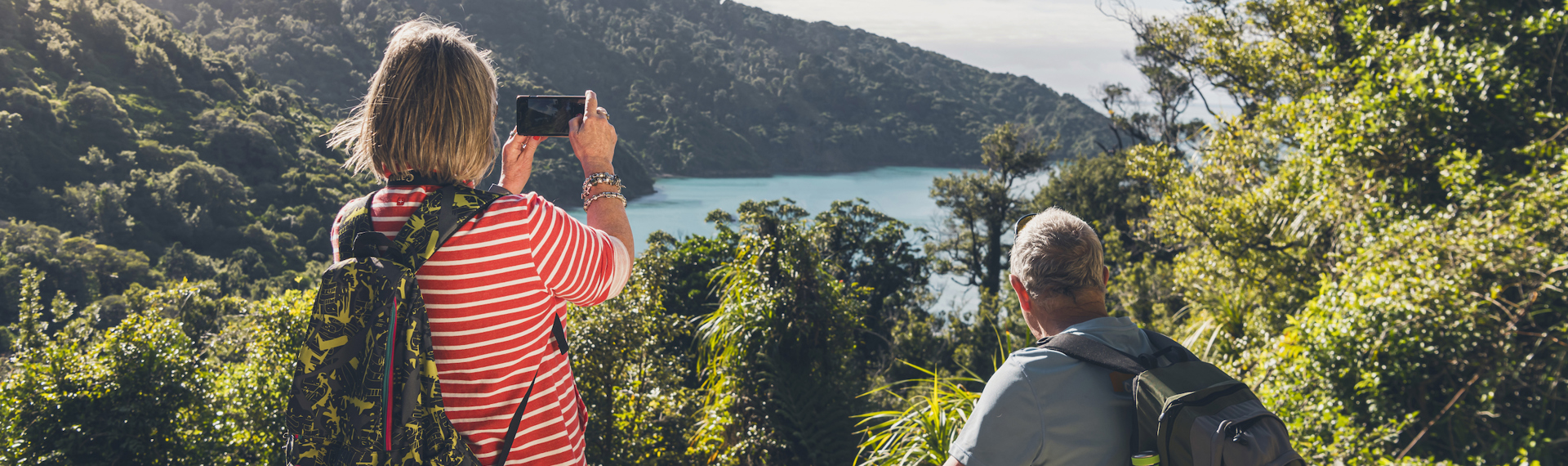 A woman takes a photo on her phone of the view across Ship Cove/Meretoto while a man sits on a bench on the northern Queen Charlotte Track in the Marlborough Sounds, New Zealand.