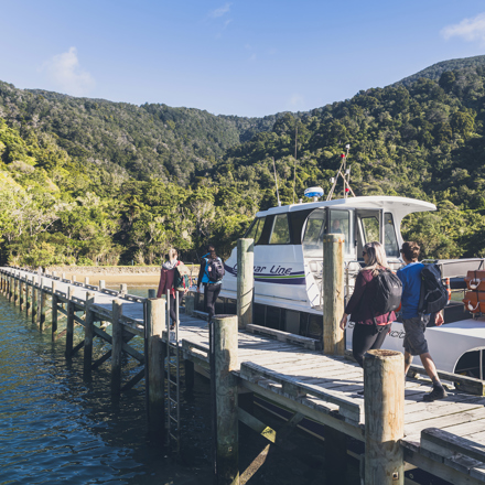 Four people walk on the jetty at Ship Cove/Meretoto next to a Cougar Line boat in the Marlborough Sounds, New Zealand.