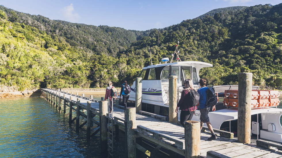 Four people walk on the jetty at Ship Cove/Meretoto next to a Cougar Line boat in the Marlborough Sounds, New Zealand.