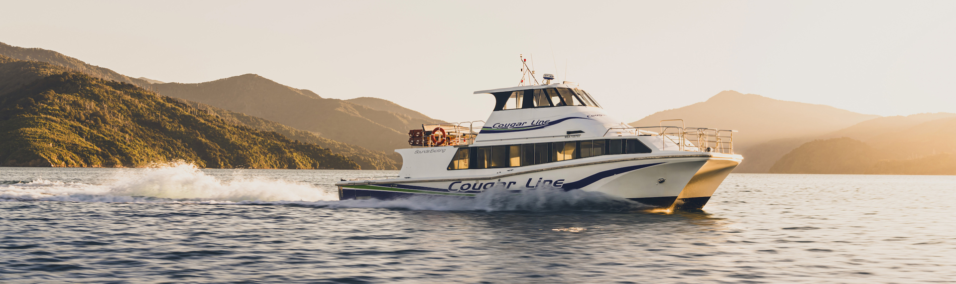 A Cougar Line boat cruises through the Marlborough Sounds under an orange sky before sunset, New Zealand.