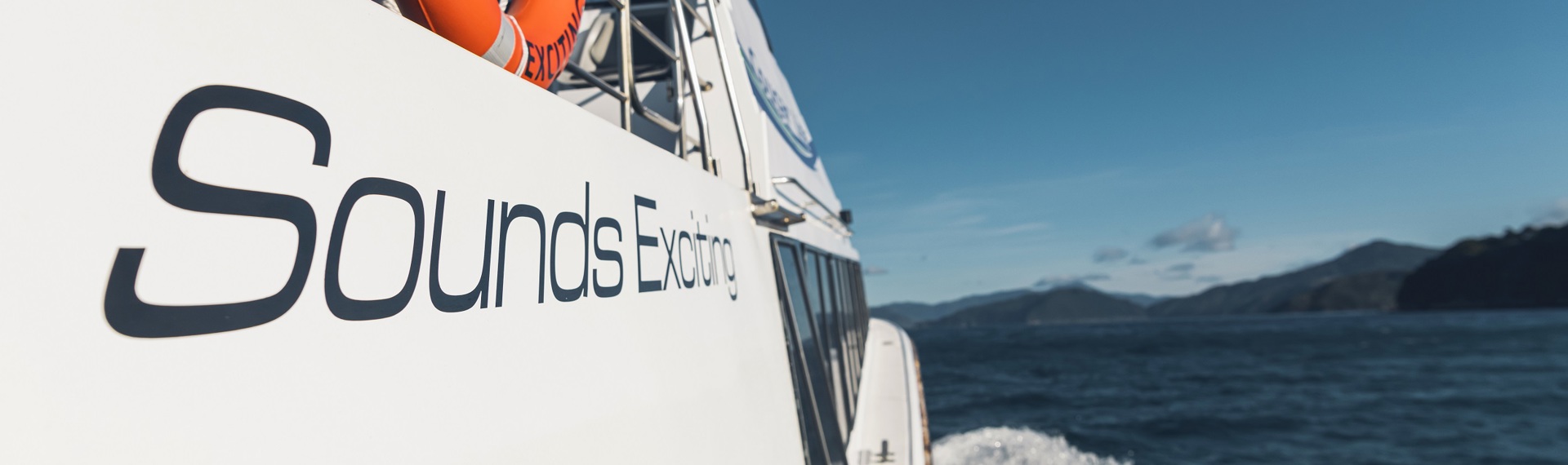 The side of Cougar Line's Sounds Exciting boat shows the boat's name, above the wake as it cruises through the Marlborough Sounds on a sunny day in New Zealand